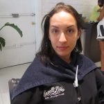 A woman sits in a chair at a salon, wearing a black robe with a towel around her shoulders