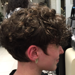 Person with short, curly brown hair, wearing gold earrings, sitting in a salon