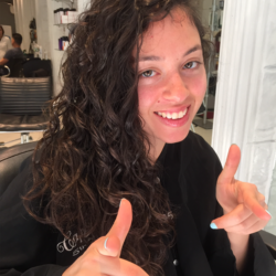 A woman with curly hair smiles while gesturing with both hands in a salon setting