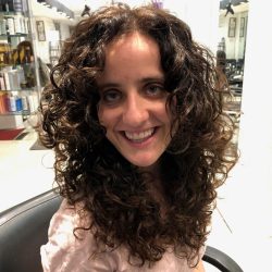 A person with long, curly hair sits in a salon chair, smiling at the camera