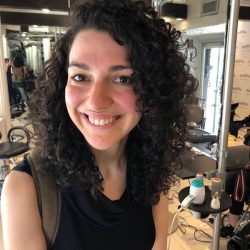 A smiling person with curly hair stands in a well-lit hair salon