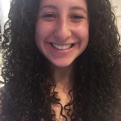 A person with long, curly dark hair is smiling and looking directly at the camera in a well-lit indoor setting