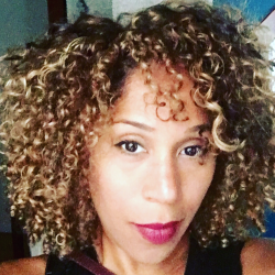 A person with curly blonde-brown hair and wearing red lipstick looks directly at the camera