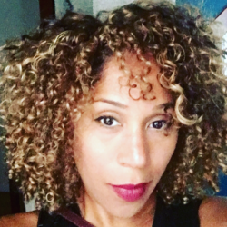 A person with curly blonde-brown hair and wearing red lipstick looks directly at the camera