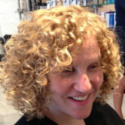 Person with curly blonde hair wearing a black shirt smiling in a brightly lit room